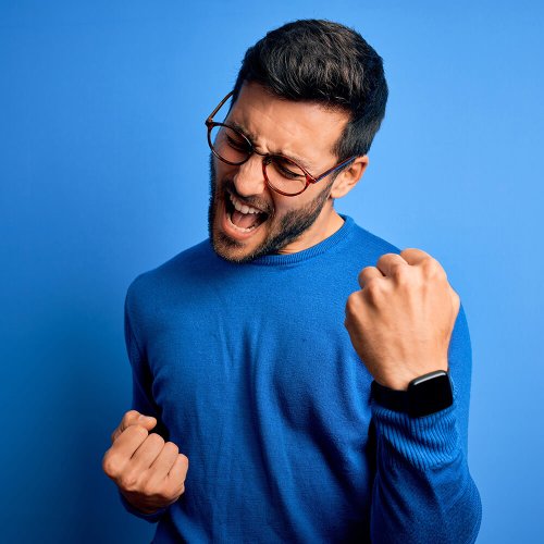 Young handsome man with beard wearing casual sweater and glasses over blue background celebrating surprised and amazed for success with arms raised and eyes closed. Winner concept.